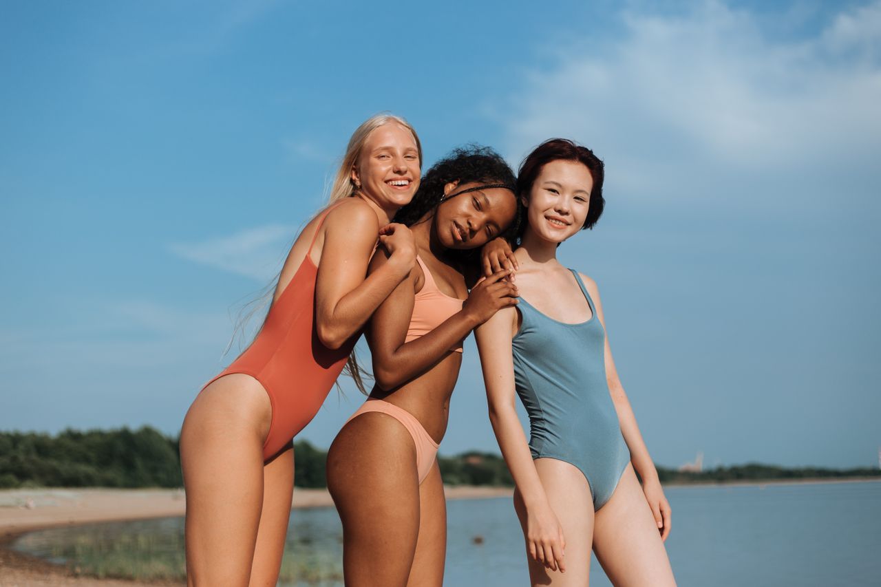 our-services-1 Three diverse teenage girls in swimwear enjoy a sunny day at the beach, smiling and embracing.