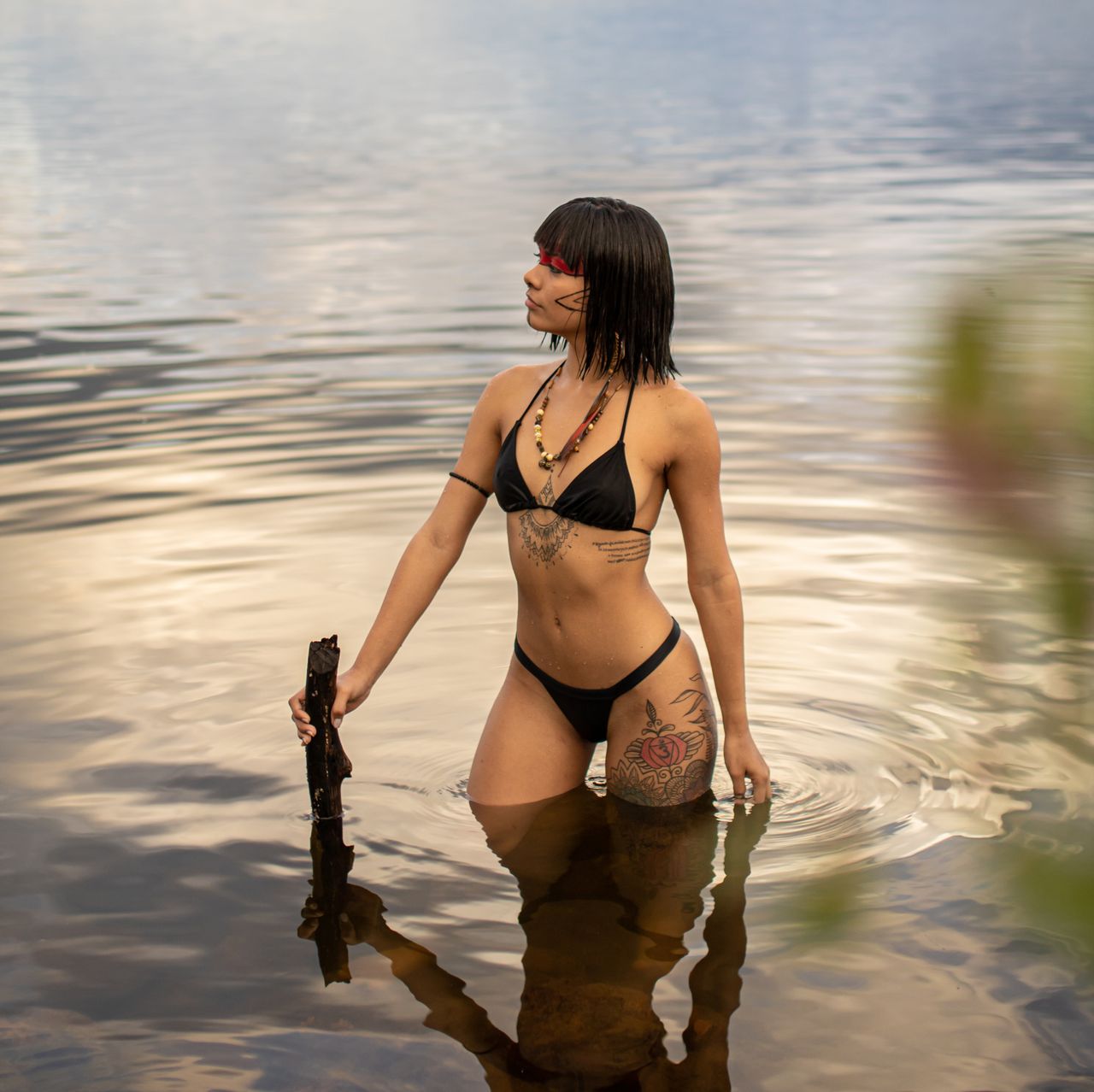 our-services-1 A woman with tattoos standing in a lake wearing a bikini, holding a stick.